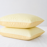 Two pale yellow pillowcases stacked on a white bed against a light gray background.