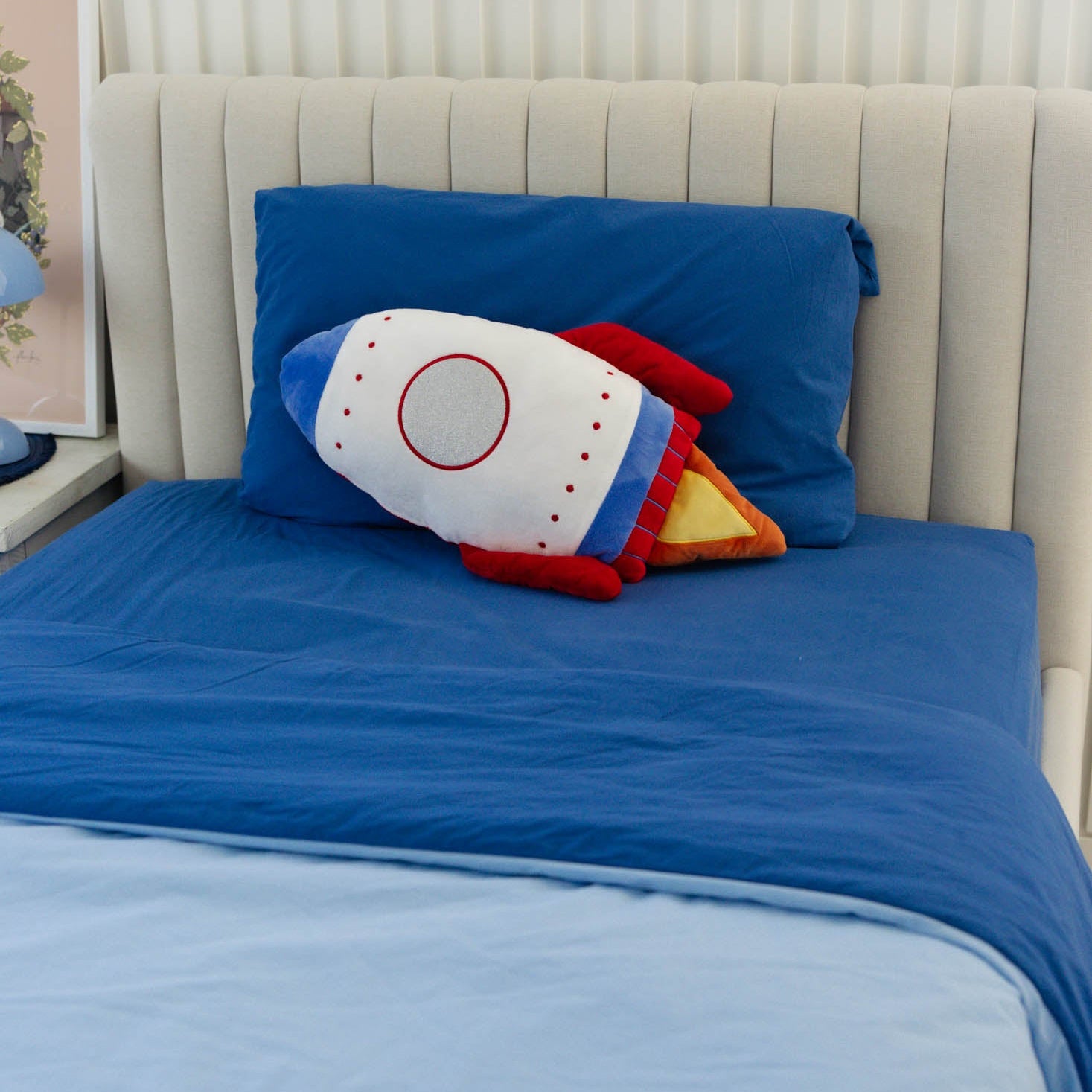 Rocket-shaped pillow on a bed with blue bedding and a white headboard.