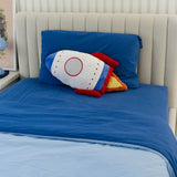 Rocket-shaped pillow on a bed with blue bedding and a white headboard.