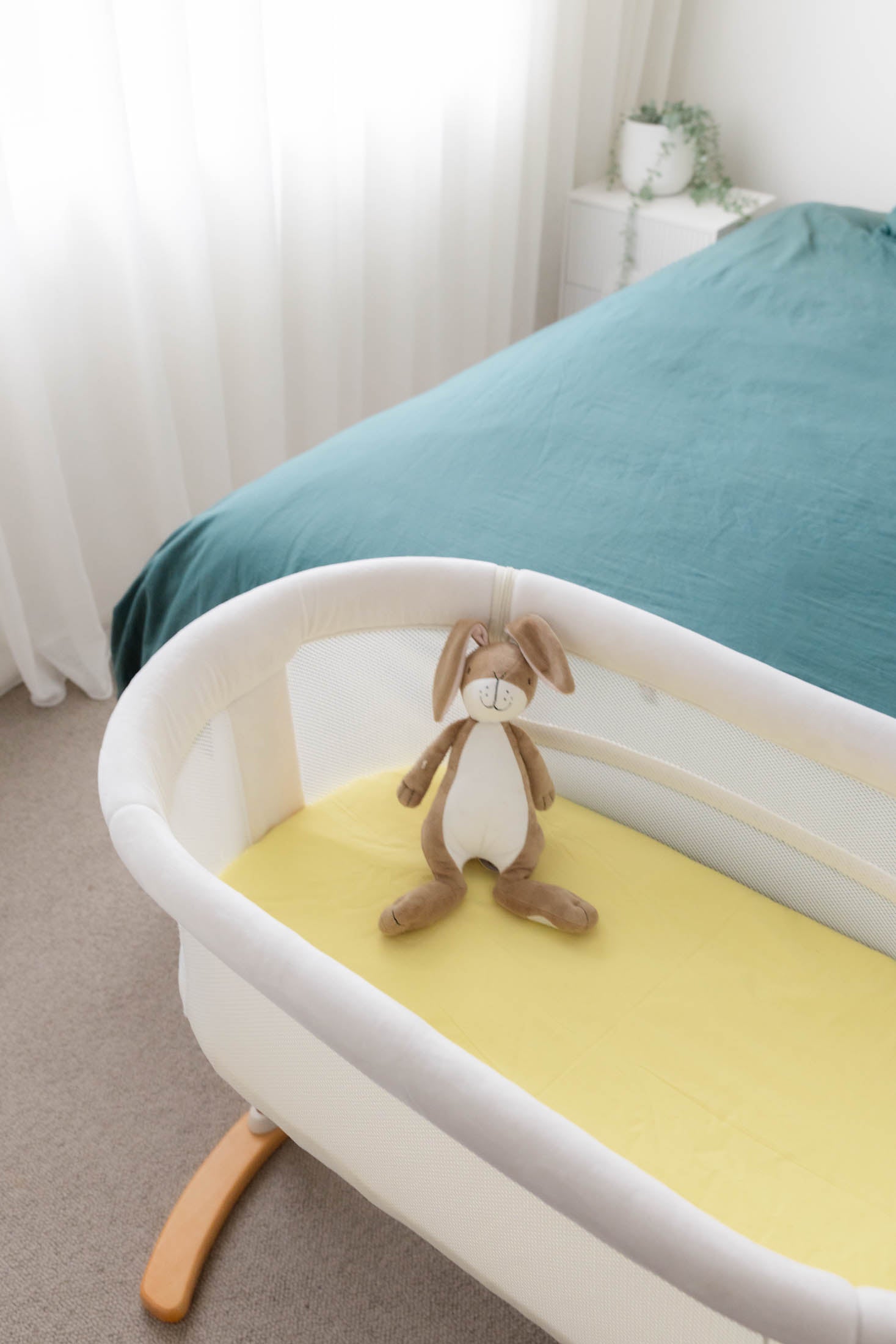 Baby crib with a yellow mattress and plush toy in a room with a bed and curtains.