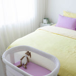 Baby bassinet next to a bed with yellow bedding and purple pillows in a bedroom setting.