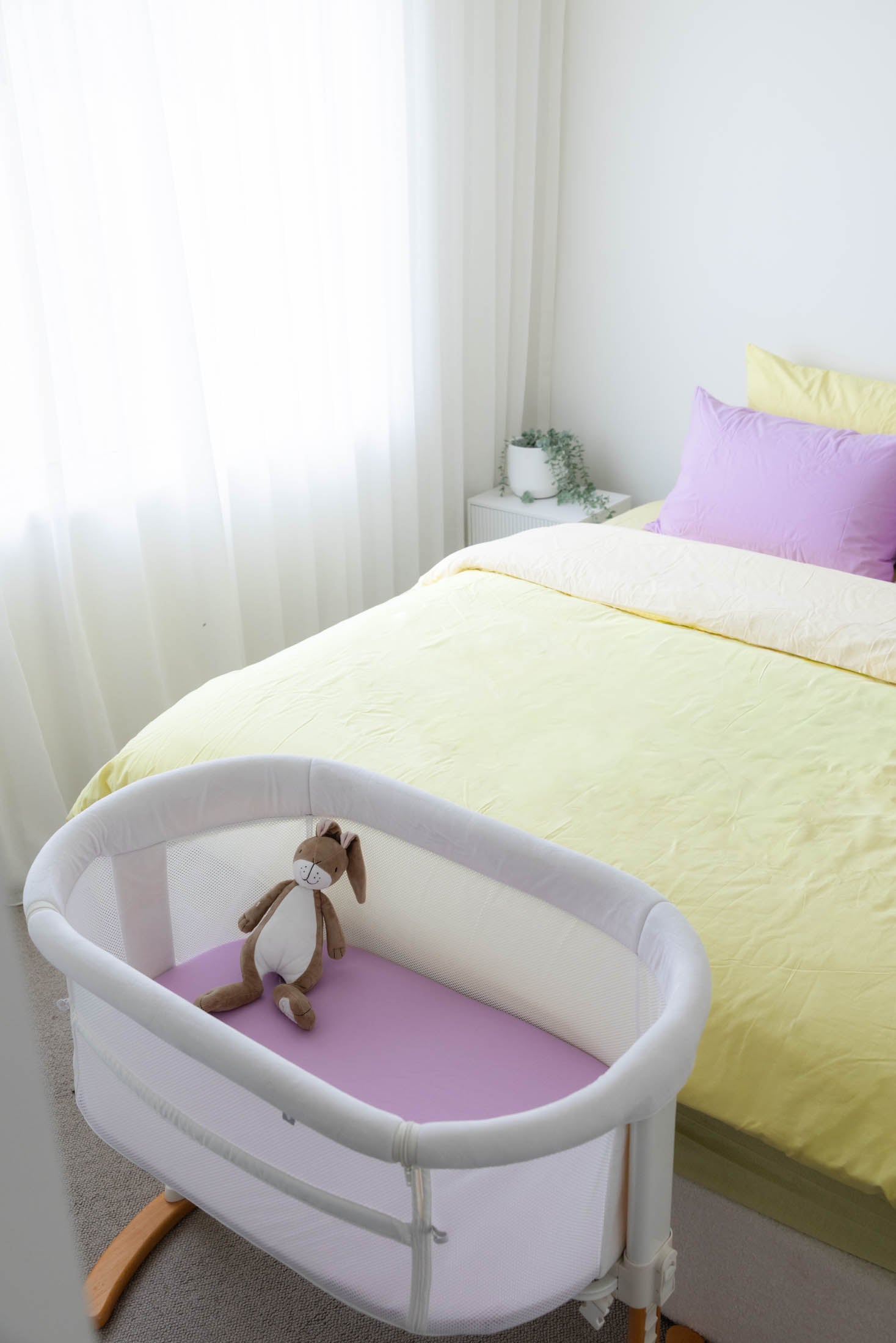 Baby bassinet next to a bed with yellow bedding and purple pillows in a bedroom setting.