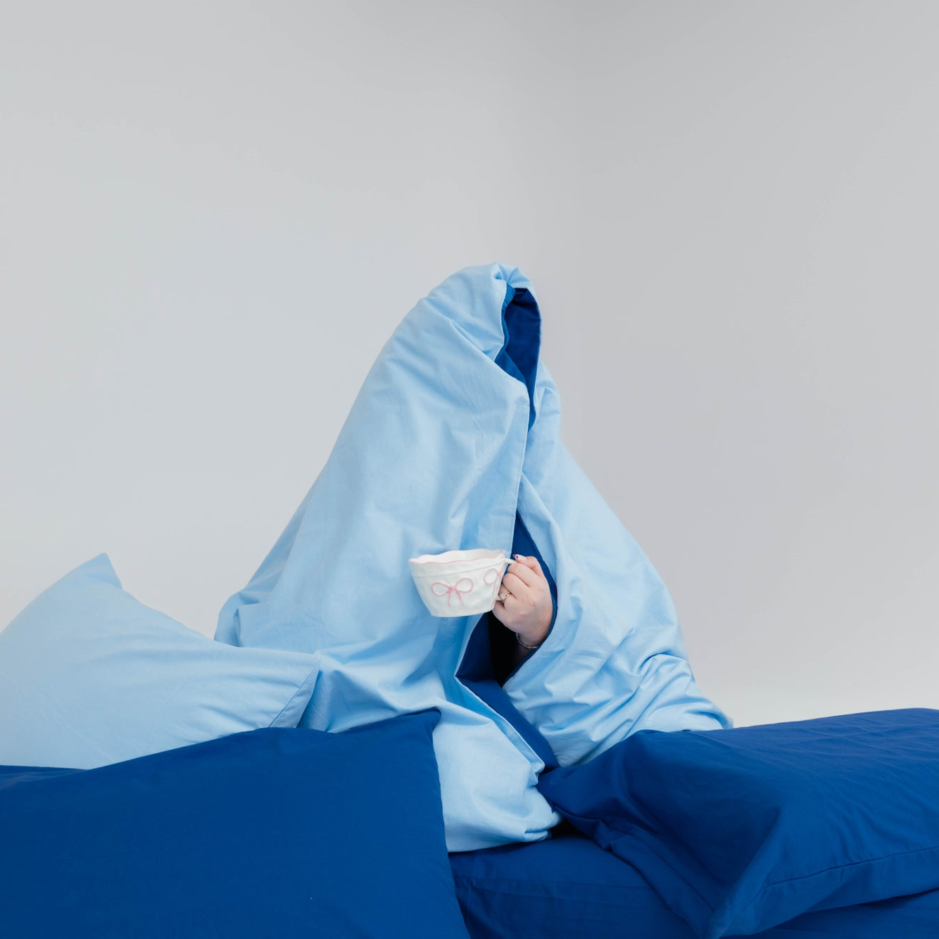 Person wrapped in a blue blanket holding a white mug with a pink design on a light gray background
