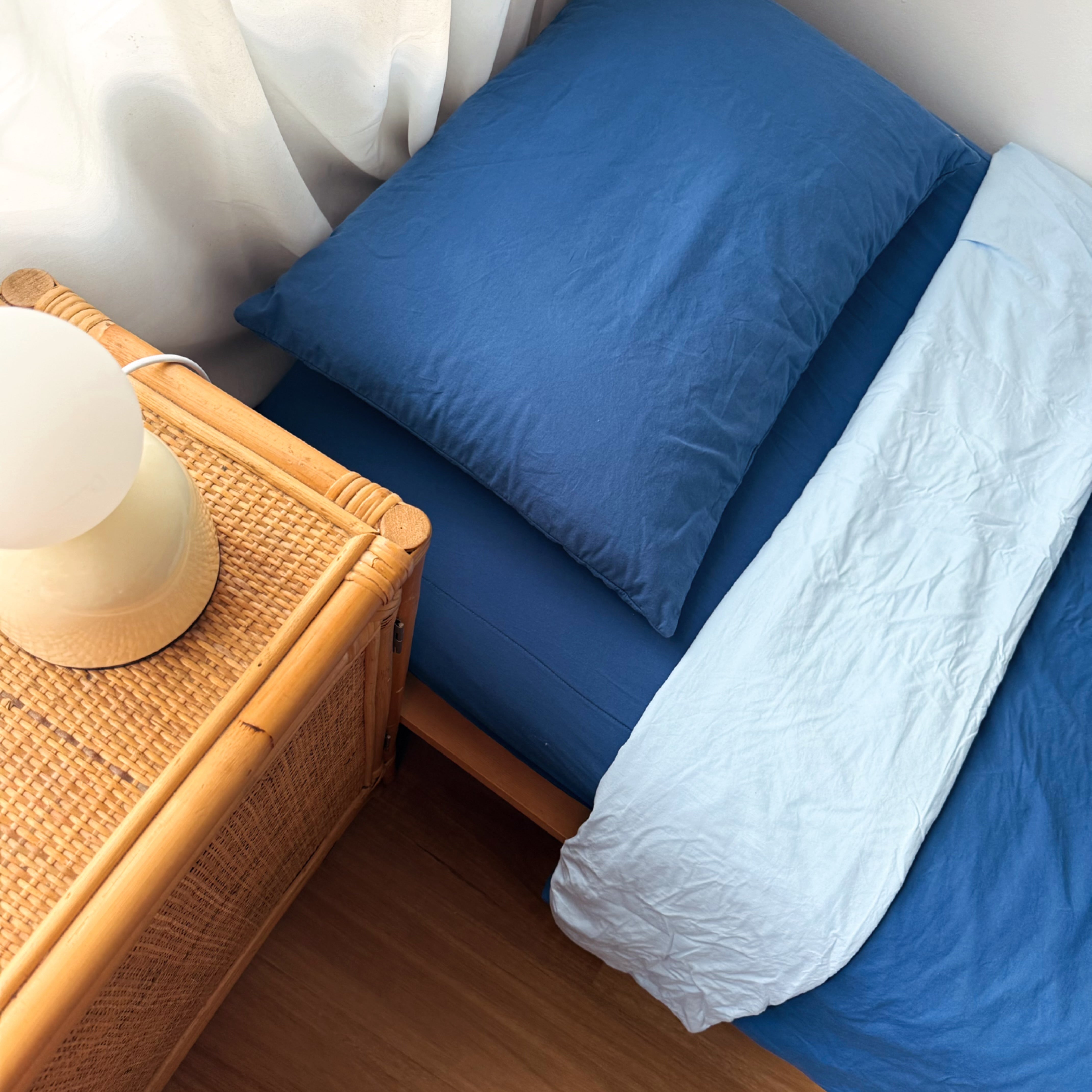 Blue bed with white blanket and blue pillow next to a wooden side table with a lamp.